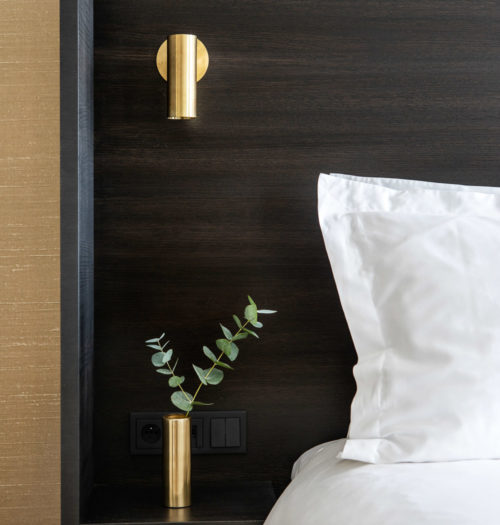 A small brass vase next to a bed with white linnen